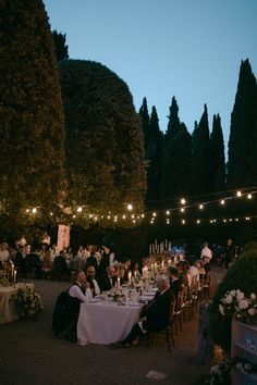 a group of people sitting around a dinner table with candles on it and trees in the background