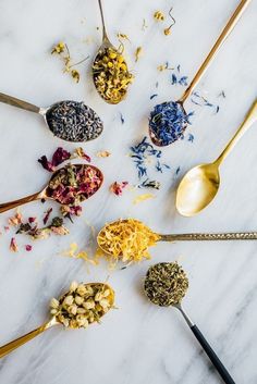 spoons filled with different types of tea on a marble counter top next to various dried flowers and herbs