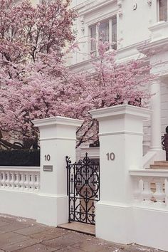 a white house with pink flowers on the trees in front of it and an iron gate