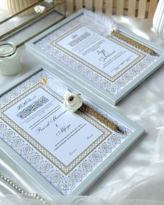 two white and gold wedding cards with a flower on them sitting on a table next to a candle