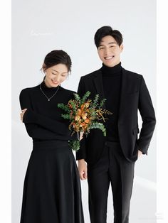 a man and woman standing next to each other in front of a white wall holding flowers