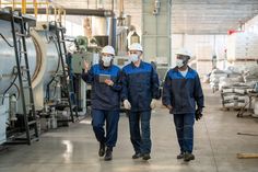 three men in blue work clothes and masks walk through an industrial area with large machines