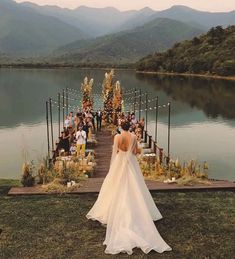 a woman in a wedding dress is standing on the edge of a dock with mountains in the background