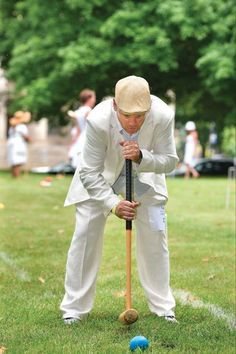 a man in white suit and hat playing croquet on grass with blue balls