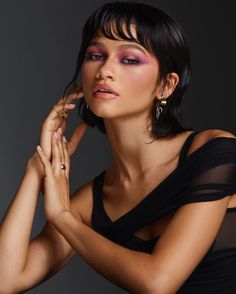 a woman posing with her hands on her chest and wearing earrings, black dress and purple eyeshadow