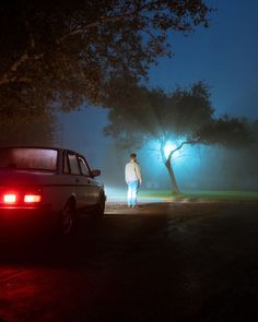 a man standing next to a car on a road at night with the lights on