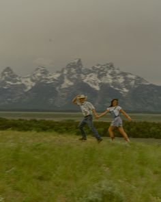 two people are running through the grass with mountains in the backgrouds behind them