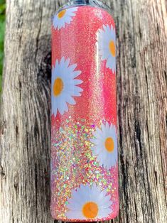 a pink and orange tumbler with white daisies on it sitting next to a tree