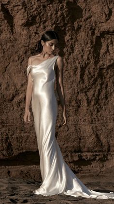 a woman standing in front of a rock wall wearing a long white dress with one shoulder