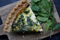 a slice of quiche with spinach and cheese on a cutting board next to greens