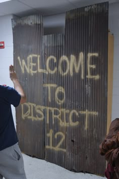 a man standing in front of a sign that reads welcome to district 12, with graffiti on it