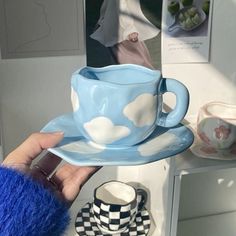 a hand holding a blue cup and saucer with clouds on the rim, in front of a mirror