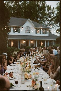 a group of people sitting at a long table with food and drinks in front of a house