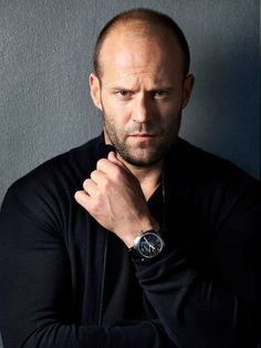 a man with a watch on his wrist standing in front of a black wall and looking at the camera