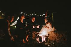 a group of people sitting around a fire pit with lights on the background at night