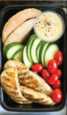 a plastic container filled with chicken, cucumbers and tomatoes next to some bread