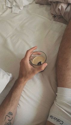 a man laying in bed holding a glass with liquid on top of his hand and another person's leg behind him