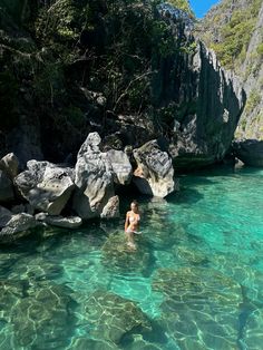 a person swimming in the clear blue water near some large rocks and trees on the shore