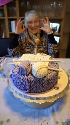 an older woman sitting at a table with a cake in front of her and waving