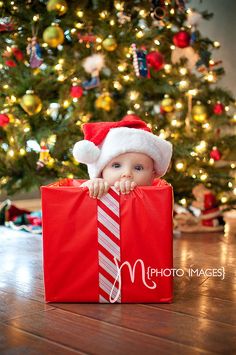 a baby wearing a santa hat sitting in a red gift bag next to a christmas tree