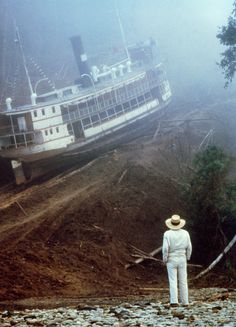 a man standing in front of a large boat on the riverbank with another boat behind him