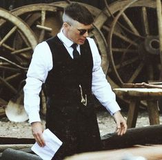 a man in black vest and tie standing next to wooden wheels