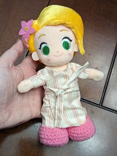 a hand holding a small doll with blonde hair and green eyes, wearing pink pants