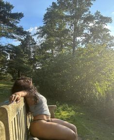 a woman sitting on top of a wooden bench next to a lush green park filled with trees