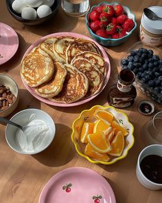 a wooden table topped with plates and bowls filled with pancakes next to fruit, nuts and other foods