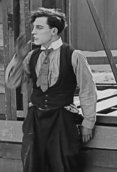 an old photo of a man standing in front of a wooden structure wearing a vest and tie