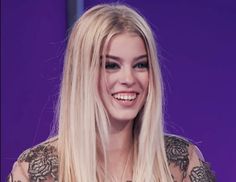 a woman with long blonde hair and tattoos on her arm smiles at the camera while sitting in front of a purple background