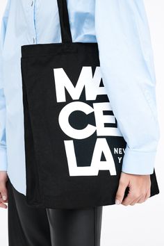 A canvas bucket tote bag in 100% European cotton, with a bottom panel for added capacity. The tote features an oversized MARCELLA NEW YORK graphic on one side, in white print on black canvas.[SPLIT] Our production team enjoys a living wage, a 40-hour work week with paid overtime, free health insurance and 20 days annual paid vacation. Canvas Tote Shoulder Bag With Logo Print, Cotton Tote Shoulder Bag With Logo Print, Casual Shoulder Bag With Logo For Daily Use, Black Cotton Shoulder Bag With Logo, Everyday Cotton Shoulder Bag With Logo Print, Daily Use Cotton Shoulder Bag With Logo Print, Canvas Bags With Logo Print For Shopping, Black Cotton Shoulder Bag With Letter Print, Everyday Cotton Shoulder Bag With Logo