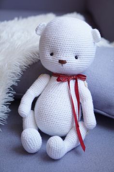 a white crocheted teddy bear sitting next to a pillow on a couch with a red ribbon around its neck