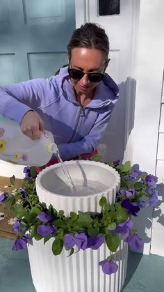 a woman is pouring water into a white container with purple flowers in it and green leaves on the rim