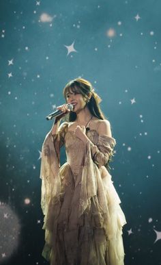 a woman in a long dress holding a microphone and standing on stage with stars behind her