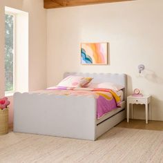 a white bed with pink and yellow pillows in a room next to a window,