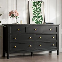 a black dresser with gold knobs in a white room next to a potted plant