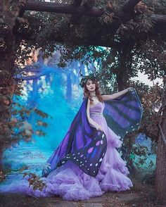 a woman in a purple dress and butterfly wings standing under a tree with her hands on her hips