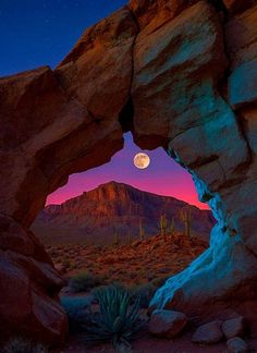 a full moon is seen through an arch in the desert, with cactus and mountains in the background