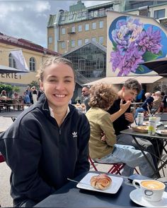 a woman sitting at an outdoor table with food and drinks in front of her smiling