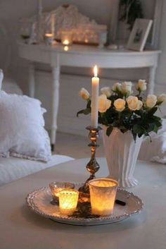 two candles are lit on a tray with flowers in the background and a vase filled with white roses