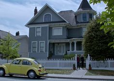 a yellow car is parked in front of a gray two story house with white picket fence