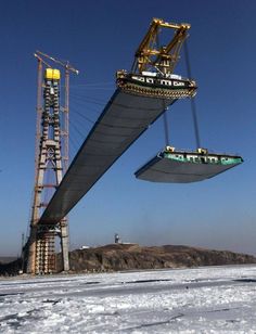 two large cranes are attached to the side of a bridge over snow covered ground with mountains in the background