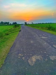 the sun is setting on an empty road in front of some green grass and trees