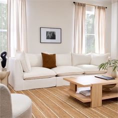 a living room with white couches and pillows on top of the sofa, coffee table in front of two windows