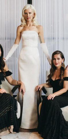 two women in dresses sitting on chairs with one woman wearing gloves and the other standing