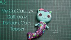 a close up of a small toy mermaid cat on a black board with words above it that read, mercat gabby's dollhouse fondant cake topper