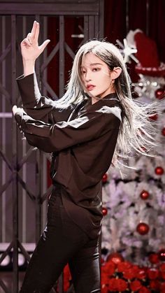 a woman with long white hair standing in front of a christmas tree and holding her hand up