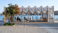 people sitting on benches near the water in front of an open air shelter with white tarps