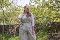 a woman standing in front of some trees wearing a gray jumpsuit and white shirt